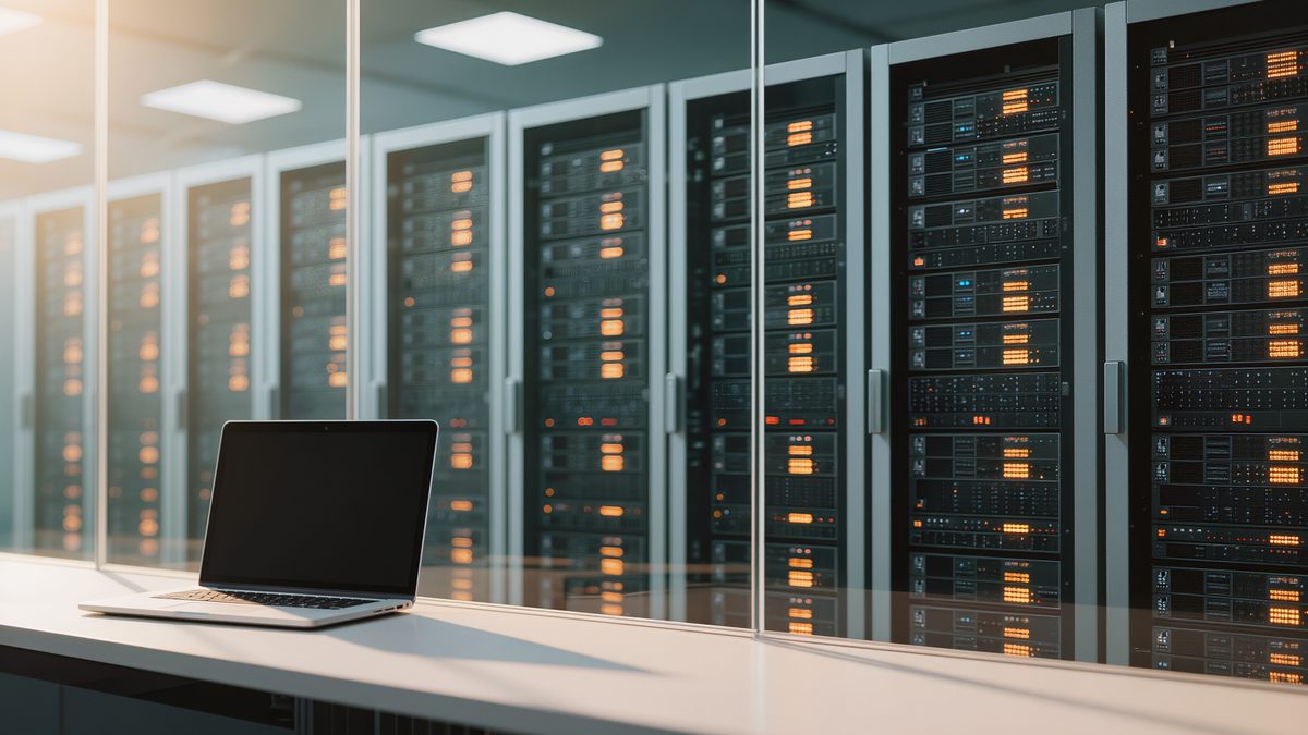 Serverraum hinter einer Glaswand mit dunklen Racks und kleinen Statuslichtern, davor ein Schreibtisch mit Laptop